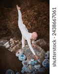 Small photo of Flexible hairless girl with alopecia in white futuristic costume put her foot up and reaches hand for surreal landscape with lot of rocks with eyes, profound symbolism of embracing individuality
