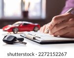 Small photo of Man signing car insurance document or lease paper. Writing signature on contract or agreement. Buying or selling new or used vehicle. Car keys on table on red car background. Warranty or guarantee.
