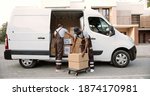 Small photo of Young handsome Caucasian delivery man giving boxes from van to African American man colleague standing on street and putting them on cart. delivering shipment. Male post office workers Courier concept