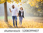 Young happy couple walking in sunny autumn garden - bearded man looking into his phone while holding hand of girlfriend - trust and privacy concept 