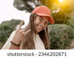 A vibrant smiling teenage girl winks, flashing a playful horn sign with one hand, reflecting the spirit of youth. She wears a pink baseball cap and looks trendy. Youth life and culture concept.