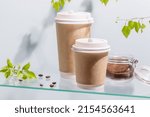 Small photo of Coffee to go. Two disposable eco-friendly cardboard cups, ground coffee in a glass jar on a glass shelf on blue background with green branches and copy space. Takeaway coffee concept. Soft focus stile