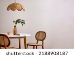 Small photo of Minimalist composition of elegant kitchen space with round table, rattan chair, vase with leaf, pedant lamp and personal accessories. Minimalist home decor. Beige wall. Template.