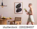 Small photo of Interior design of aesthetic and elegant room with woman in dress, mock up poster frame, round table, vegetables, rattan chair and personal accessories. Stylish home decor. Template.