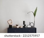 Small photo of Minimalist composition of elegant and outstanding space with navy commode, green leaves in vase books, black clock, sculpture and personal accessories. Home decor. Template. Copy space.