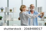 Small photo of Caucasian old senior elderly happy grandparents lover married couple standing smiling look at each other face listening to slow waltz music dancing together in living room at home in front bookshelf.