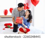 Millennial Asian young romantic lover couple male boyfriend giving present wrapped gift box surprised to female girlfriend on bed decorated with heart shape balloon on valentine day anniversary.