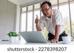 Small photo of Asian old senior happy friendly male businessman in business outfit with glasses waving hand smiling say hello greeting with customer via video call conference from laptop computer in company office.