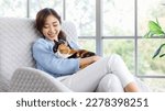 Small photo of Millennial Asian young female owner sitting on cozy armchair smiling holding hugging cuddling showing love to cute fat tabby tricolor short hair little domestic furry purebred pussycat in arms.
