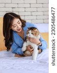 Small photo of Millennial Asian young kindly cheerful female owner sitting on bed holding hugging cuddling showing love with long hair cute little domestic kitten furry pussycat pet friend on blanket