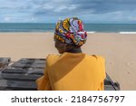 Small photo of Ghanaian woman on the beach wearing African colorful headdress and sitting on a wooden bench in keta village