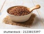 Beige ceramic bowl full of raw wholegrain buckwheat groats and spoon on a white wooden table. Cooking buckwheat porridge. Concepts of gluten free diet and vegetarian food. Close-up.