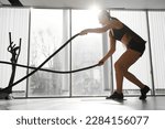 Small photo of Athletic Female in a Gym Exercises with Battle Ropes During Her Fitness Workout High-Intensity Interval Training. She's Muscular and Sweaty.