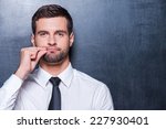 Keep silence! Handsome young man in formalwear showing a sign of closing mouth and looking at camera while standing against blackboard