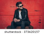 Gorgeous and stylish. Handsome young man in sunglasses keeping hands clasped and looking away while sitting on wooden chest against red background