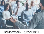 Business coach. Rear view of man gesturing with hand while standing against defocused group of people sitting at the chairs in front of him 