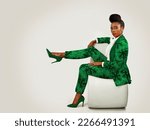 Small photo of Confident African business woman wearing an African print suit and heels with a sophisticated hairstyle sitting in a clean space with her leg propped up.