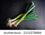 Overhead View of Whole Leeks on a Dark Background: Three leeks with leaves and roots on a stone countertop
