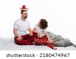Small photo of Family in plaid sleepwears sitting on bed on white studio. Daddy holds book and cup of coffee on heady. Single dad and сurly short-haired daughter in red pajamas having fun and leisure time together