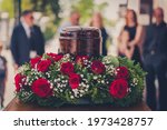 Funerary urn with ashes of dead and flowers at funeral. Burial urn decorated with flowers and people mourning in background at memorial service, sad and grieving last farewell to deceased person.