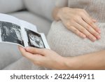 Close-up of a pregnant woman sitting in the living room at home looking at the ultrasound image of her own baby. Happy pregnancy woman during prenatal ultrasound at her home