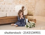 Small photo of Smiling little curly hair girl with a bouquet of jasmine flowers sits by the sofa in cozy autumn living room. Scandinavian interior. Easter.ation. Mothers Day. Child with a bouquet of flowers for mom