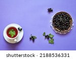 Ripe garden freshly picked blackcurrant on saucer and cup of vitamin hot tea with currant leaf on a purple background. Free space for text. Organic Vitamin Seasonal Products. Flat lay, copy space