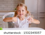 baby blonde shows thumbs up with a glass of water in her hand. Child girl holding a glass of water. Child recommend drinking water. Good healthy habit for children. Healthcare concept