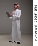 Small photo of A saudi character wearing thob standing working on tablet on withe background.