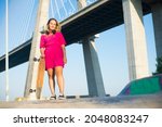 Portrait of smiling young woman holding longboard. Beautiful girl in headphones and dress with skateboard standing near bridge. Sport, hobby, active lifestyle concept