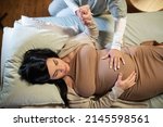 Small photo of Top view of smiling pregnant woman and midwife at home. Woman in casual clothes lying on bed, midwife holding hand. Pregnancy, medicine, home birth concept