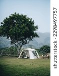 Small photo of A white tent under a tree on a green grass with mountain on the background, with camping gears and chairs