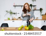 Small photo of A woman with red hair is standing in her kitchen smiling and cooking celery juice with a juicer