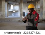 African american workman in yellow hard hat stands at construction site, holds in hands smartphone and dials number