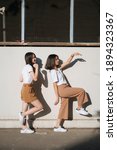 Small photo of Two girls in white t-shirt and brown pants do the silly act together in the sunlight at the wall.