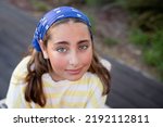 Outdoor portrait of beautiful happy teen girl with rosy cheeks. Young woman with beautiful big green eyes. Smiling face. Eye contact. Stylish image, blue bandana. Sunset. Beauty, embarrassment concept