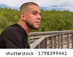 Small photo of Outdoors leaning on the boardwalk rail, he looks across the way with a staring gaze. A scared face tough guy with a military buzz cut leans on the wood rail with a focused look.