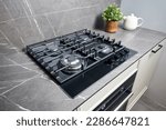 Modern hob gas stove made of tempered black glass using natural gas or propane for cooking products on light countertop in kitchen interior with oven tea pot and flowers.