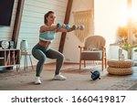 Beautiful young athletic girl in leggings and top crouches with dumbbells at home. Sport, healthy lifestyle.
