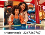 A cute African-American child with afro curls with her mother playing air hockey at an amusement park and carousel on her day off in the evening.