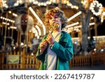 A cute caucasian boy with blonde curly hair eating a colorful lollipop standing against the background of a carousel with horses in the evening at an amusement park or circus.