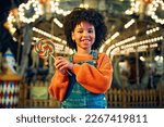 A cute African-American girl with an Afro hairstyle eats a colorful lollipop standing against the background of a carousel with horses in the evening at an amusement park or circus.
