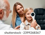 Small photo of Cute little girl closing one eye during an ophthalmologist consultation while sitting on her mother's lap. A child points to an eye chart during an eye test.