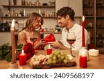 Happy Valentine's Day. A couple with glasses of wine by candlelight sitting in the kitchen at the table, romantically spending the evening together. A man giving his beloved a heart-shaped gift box.