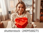 Happy Valentine's Day. Happy smiling woman holding red heart shaped gift box with bow in living room at home.