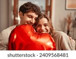 Happy Valentine's Day. Young couple in love holding a heart-shaped balloon, hiding behind it while sitting on the sofa in the living room at home. Romantic evening together.