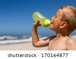 Small photo of Upper body shot of thirsty boy who is drinking water from his bright green water bottle. Boy is standing at beach while the sun is shining without mercy. Ocean waves and blue sky in the background.