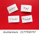 Small photo of Torn paper pieces on red background with bully words UGLY STUPID WEIRDO IDIOT, concept of someone who hurts or frightens victims by degrade or demean in some way to feel powerful or stronger