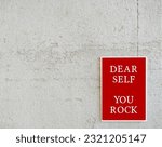 Small photo of Red poster on copy space white wall with text message DEAR SELF YOU ROCK, positive self talk to raise self esteem, daily beautiful affirmation to remind strength and ability to overcome difficulties