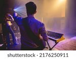 Small photo of Young latin bassist and keyboardist with shirt and glasses, and cream bass and vintage keyboard playing live in a concert under colorful lights